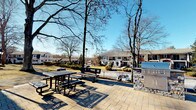 The Fire Pit area is a well-appointed outdoor amenity space designed for community gathering and relaxation. The foundation of this space is a generously sized stone-tile patio, laid in a neutral palette that complements the natural surroundings. The area is framed by the building's striking exterior, characterized by bold terracotta-orange architectural panels, large floor-to-ceiling windows, and clean white trim — creating a modern yet welcoming aesthetic.

The outdoor space is thoughtfully furnished with metal picnic tables and benches, providing ample seating for casual dining or socializing. A stone-clad built-in grill station serves as a functional centerpiece, ideal for outdoor cooking. Curved pathways extend across the patio, connecting to additional outdoor lounge seating areas outfitted with cushioned sofas and chairs, perfect for relaxed conversation.

Beyond the patio, the landscape opens to views of mature trees, manicured lawns, and neighboring residential buildings, offering a tranquil, park-like backdrop. White market umbrellas near what appears to be a pool area are visible in the distance, suggesting a broader outdoor amenity complex. The space benefits from abundant natural light and open skies, making it an ideal setting for outdoor gatherings throughout the seasons. Large glass entry doors provide seamless indoor-outdoor access directly from the building.