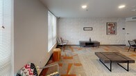Located at 13280 Linden Ave N in Seattle, WA, this single-floor community space is thoughtfully designed to support a variety of social and functional activities. The layout is open and cohesive, connecting distinct functional zones in a seamless flow.

The kitchen area is outfitted with warm wood cabinetry, dark countertops, stainless steel appliances including a full-size refrigerator and microwave, and a central island with a sleek gray surface. Light tile flooring and recessed lighting give the space a clean, modern aesthetic. An accessible restroom is conveniently located adjacent to the kitchen.

The lounge area features a distinctive, vibrant geometric-patterned carpet with warm orange and neutral tones, providing a welcoming atmosphere. Large windows along the exterior wall allow ample natural light to fill the room. Seating is flexible, complemented by a low media console and wall-mounted display.

A dedicated dining area accommodates group gatherings around a solid wood table with metal wire chairs. Framed artwork on the walls adds a curated, finished touch to the space. The overall design balances contemporary finishes with functional, community-oriented features, making this space well-suited for shared use and social interaction.