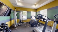 This fitness center at 11400 Edgemere Blvd in El Paso, Texas, is a compact yet well-equipped exercise room designed to support a variety of workout routines. The space is immediately distinguished by its bold, horizontally striped walls in yellow, sage green, and gray, creating an energetic and motivating atmosphere. A tray ceiling with recessed lighting adds a sense of height and polish to the room.

The exercise room is outfitted with a strong selection of cardio equipment, including treadmills, an elliptical, a stair climber, and a recumbent bike — several of which are Cybex brand machines. A multi-station cable machine and a weight bench provide options for strength training, while a rack of dumbbells and medicine balls caters to free-weight workouts. A wall-mounted flat-screen television adds entertainment value during exercise sessions.

Natural light filters into the space through multiple windows fitted with blinds, offering outdoor views of the surrounding greenery. A wall-mounted mirror further enhances the sense of openness. The durable rubber flooring is well-suited for a high-activity environment. The room's efficient layout maximizes the use of the available floor area, making it a practical and functional fitness facility for residents of the property.