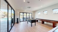 Located at 2420 Cardinal Drive in San Diego, CA, this contemporary game room is a well-appointed recreational amenity space designed for social entertainment. The room features light hardwood-style flooring and crisp white walls that give the space a clean, modern aesthetic. Recessed lighting illuminates the interior, complemented by abundant natural light streaming through large, black-framed floor-to-ceiling sliding glass doors that span the length of one wall.

The game room is equipped with a full lineup of recreational equipment, including a foosball table, an air hockey table, and a shuffleboard table, offering a variety of activities for guests. A wall-mounted flat-screen television serves as a central entertainment focal point, while tall bar-height tables with swivel stools provide casual seating throughout.

The striking black-framed sliding glass doors create a seamless indoor-outdoor connection, opening directly onto an outdoor patio area furnished with dining tables, chairs, and a built-in grill — ideal for gatherings and alfresco entertainment. Additional accent windows along the upper walls bring in supplementary natural light while maintaining privacy. The overall design balances modern minimalism with a welcoming, activity-focused atmosphere suited for community recreation.