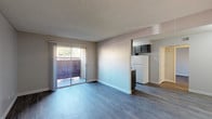 This single-floor space features an open-concept living area and a functional galley kitchen, connected in a practical layout. The living area is finished with durable laminate wood-style flooring in a cool gray tone, complemented by neutral beige walls and a textured ceiling. A large sliding glass door floods the space with natural light and opens to a private outdoor patio, offering a seamless indoor-outdoor connection. The open passage to the kitchen is partially defined by a half-wall, maintaining a sense of openness throughout.

The galley kitchen is efficiently designed with dark cabinetry lining both sides, providing ample storage. Granite-look laminate countertops offer functional workspace, while the kitchen is equipped with a white electric range, a full-size refrigerator, and a dishwasher. A window at the far end of the kitchen brings in additional natural light and outdoor views. The matching laminate flooring continues throughout, tying the spaces together cohesively. A door visible near the kitchen suggests access to additional rooms or utility areas. Overall, the space offers a clean, functional, and well-lit environment.