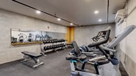 Located at 29 Crown Street in New Haven, CT, this fitness center offers a well-equipped workout facility across a single floor. The main exercise area features durable rubber-tile flooring and textured concrete-style walls, creating a functional and industrial aesthetic. Recessed lighting throughout ensures a well-lit environment for workouts at any time of day. A large wall-mounted mirror spans one side of the weight area, enhancing the sense of space and allowing users to monitor their form. The facility is comprehensively equipped with a multi-tier dumbbell rack holding a full range of free weights, multiple workout benches, Precor treadmills, elliptical trainers, a stationary bike, a rowing machine, and a Cybex multi-function cable and strength machine. Exposed overhead piping and ductwork add to the space's industrial character, while a wall-mounted air conditioning unit provides climate control. A separate, accessible bathroom is also included, featuring light hardwood-style flooring, clean white walls, ADA-compliant grab bars, a wall-mounted sink, and a warm wood-framed mirror, making the facility accommodating for all users.