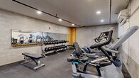Located at 29 Crown Street in New Haven, CT, this fitness center offers a well-equipped workout facility across a single floor. The main exercise area features durable rubber-tile flooring and textured concrete-style walls, creating a functional and industrial aesthetic. Recessed lighting throughout ensures a well-lit environment for workouts at any time of day. A large wall-mounted mirror spans one side of the weight area, enhancing the sense of space and allowing users to monitor their form. The facility is comprehensively equipped with a multi-tier dumbbell rack holding a full range of free weights, multiple workout benches, Precor treadmills, elliptical trainers, a stationary bike, a rowing machine, and a Cybex multi-function cable and strength machine. Exposed overhead piping and ductwork add to the space's industrial character, while a wall-mounted air conditioning unit provides climate control. A separate, accessible bathroom is also included, featuring light hardwood-style flooring, clean white walls, ADA-compliant grab bars, a wall-mounted sink, and a warm wood-framed mirror, making the facility accommodating for all users.