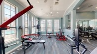 This fitness center offers a single, well-appointed exercise room designed to support a thorough strength training regimen. The space is characterized by its elevated ceilings adorned with crown molding and ceiling fans, contributing to an open, airy atmosphere. Soft teal-painted walls paired with neutral, multi-toned carpet tiles create a clean and focused environment conducive to exercise.

Natural light flows generously through multiple large windows along the perimeter walls, providing views of the surrounding outdoor area and keeping the space bright and inviting. Recessed lighting supplements the natural light for consistent illumination throughout.

The exercise room is comprehensively stocked with a wide variety of strength training equipment, including barbells, weight benches, squat racks, dumbbell racks, kettlebells, cable machines, and Titan Fitness branded equipment. Floor-to-ceiling mirrors line one wall, visually expanding the space and allowing users to monitor their form during workouts. The layout is thoughtfully organized to accommodate multiple users simultaneously, with clear zones for free weights and machine-based exercises. White-trimmed arched entryways add an architectural detail that elevates the overall design of the space.