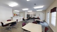 This single-floor multipurpose room is designed to accommodate a variety of activities, from meetings and classes to social gatherings. The space is furnished with several light-colored rectangular tables paired with burgundy and black stackable chairs, arranged in a flexible layout that can be easily reconfigured to suit different needs. Neutral-toned carpet covers the floor, contributing to a quiet and comfortable atmosphere. Overhead, a series of rectangular fluorescent light fixtures provide consistent, even illumination throughout the room. Along one wall, a white reception or service counter is visible, suggesting a check-in or administrative function. A dark storage or display cabinet stands nearby, alongside a low credenza running beneath a pair of framed artwork pieces that add a touch of visual interest to the walls. Colorful canvas artwork near the counter brightens the space further. A standard door on the far wall provides access to an adjacent area. The overall design is practical and functional, prioritizing usability and accessibility within a clean, well-maintained environment.
