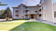 This residential building presents a welcoming exterior characterized by neutral-toned lap siding, structured across multiple stories with a clean, functional design. The building's facade is punctuated by numerous windows that allow for generous natural light throughout its units, while private balconies on each level offer residents their own outdoor retreat with views of the surrounding grounds.

The covered main entrance is a notable architectural feature, highlighted by a gabled canopy supported by stone-clad columns that lend a warm, inviting character to the building's entry point. A concrete walkway leads directly to the glass-paneled front doors, framed neatly by the stone and wood detailing.

The building is set within well-maintained landscaping, featuring an expansive open lawn that contributes to a serene and open atmosphere. Mature trees and greenery border the property, enhancing privacy and adding natural beauty to the setting. The single-floor unit layout offers a straightforward, accessible living experience within a multi-story building context.

Overall, the building combines practical construction with thoughtful exterior detailing, creating a comfortable and visually appealing residential environment suited for everyday living.