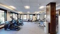 Located at 460 Wacouta St in Saint Paul, Minnesota, this fitness center offers a well-appointed exercise facility designed to support a comprehensive workout experience. The space is organized across a single floor and is divided into two functional zones, providing ample room for both cardio and strength training activities.

The main exercise area is lined with large, dark-trimmed windows along multiple walls, flooding the space with natural light and offering views of the surrounding outdoor environment. The layout accommodates a robust selection of cardio equipment, including treadmills, elliptical trainers, and stationary bikes, all positioned to take advantage of the natural light and exterior views. Wall-mounted televisions are strategically placed throughout for convenience during workouts.

The strength training section features a multi-function cable machine and various resistance equipment. Full-length mirrors are integrated into the design, enhancing the sense of openness while providing functional utility for form monitoring. Dark wood architectural columns add a refined, upscale aesthetic that contrasts with the neutral-toned carpet flooring and drop-panel ceiling with recessed lighting.

A separate room, accessible through glass-paneled doors, provides additional training space equipped with free weights and exercise balls, offering a more private environment. A water fountain is conveniently located near the entry corridor. The overall ambiance is clean, modern, and professional, reflecting a thoughtfully designed facility.
