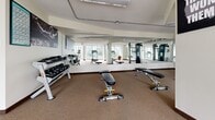 Located at 102-120 Agnes Street in New Westminster, BC, this fitness facility offers a well-appointed and thoughtfully organized workout space across a single floor. The gym is designed to accommodate a wide range of exercise activities, making it suitable for both cardio training and strength work.

Large floor-to-ceiling windows line the perimeter of the cardio zone, flooding the space with natural light and providing pleasant views of the surrounding greenery and mature trees. This area houses multiple treadmills and stationary bikes, creating a bright and motivating environment for aerobic exercise.

Full-length mirrors span entire wall sections throughout the gym, visually expanding the space and providing useful feedback during workouts. A motivational wall decal reinforces an active atmosphere. The strength training area is well-stocked with a complete rack of dumbbells and multiple adjustable weight benches, supported by a cable machine for versatile resistance training.

A dedicated functional training and stretching zone features a raised platform area, exercise mats, stability balls, and open floor space — ideal for group classes, yoga, or dynamic movement exercises. A wall-mounted split air conditioning unit ensures year-round climate comfort. Neutral carpet flooring and a clean, white interior palette give the gym a practical yet welcoming ambiance throughout.