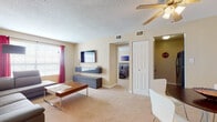 This 1-bedroom, 1-bathroom unit offers a practical and well-organized single-floor layout designed for comfortable everyday living. The carpeted living room serves as the central gathering space, featuring a wall-mounted flat-screen TV, floating media console, and a large sectional sofa paired with a wooden coffee table. A generous window dressed with bold red curtains allows natural light to fill the room, while a ceiling fan overhead provides year-round comfort. Adjacent to the living area is a compact dining nook with a small round table and modern chairs, positioned near the front entry with a closet for additional storage. The bedroom is a peaceful retreat, offering a full closet with built-in shelving and a dresser, complemented by a window with blinds and curtain panels that regulate light. Neutral-toned walls and plush carpeting carry throughout the private spaces, creating a cohesive and relaxed ambiance. The full bathroom features tile flooring, a dark wood vanity with a white countertop, a wall mirror with overhead lighting, and a shower-tub combination enclosed by a curtain. Overall, this unit delivers a clean, functional living experience with a neutral palette and thoughtful use of its available space.
