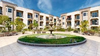 Located at 14105 Chadron Ave in Hawthorne, California, this outdoor courtyard space showcases a distinctive Mediterranean-inspired architectural design. At the heart of the courtyard stands a classic multi-tiered stone fountain, encircled by a lush green lawn and curved stone retaining borders that define the central garden feature. The surrounding plaza is finished with large-format beige tiles, providing ample open space ideal for gathering and leisure.

The courtyard is enclosed by a multi-story residential building with warm stucco exteriors in complementary earth tones of beige and terracotta. Each floor features private balconies with black iron railings, lending a refined, cohesive aesthetic to the façade. Arched architectural details, decorative medallions, and varied rooflines add visual character throughout.

Thoughtfully landscaped raised planters line the perimeter, filled with a variety of shrubs, flowering plants, palm trees, and mature shade trees, creating a lush, resort-like ambiance. Seating areas with wooden benches and umbrella-shaded patio tables are distributed throughout the space, offering comfortable spots for residents to relax outdoors. The open-air design allows for abundant natural light and a welcoming sense of community within this well-maintained Southern California property.