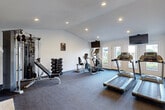 Located at 6075 Grelot Rd in Mobile, Alabama, this single-story fitness center is thoughtfully divided into two distinct workout zones, each designed to support a different style of training. The main cardio and strength room features durable carpet flooring and is equipped with Matrix treadmills, an elliptical, a stationary bike, and a multi-station cable machine accompanied by a full rack of dumbbells. Recessed lighting and vaulted ceilings lend the space an open, airy feel, while a wall-mounted flat-screen TV and French doors with views of the outdoors enhance the workout environment. A standout architectural feature of this room is a classic white-painted brick fireplace, adding a unique and warm aesthetic contrast to the fitness setting. The adjacent functional training studio offers a smooth hardwood-style floor ideal for dynamic movement and flexibility work. This room is outfitted with a multi-tier functional training rack holding medicine balls, kettlebells, and TRX suspension equipment, along with a set of stability balls and a BOSU trainer. A large wall-mounted TV and natural light from multiple windows complete the space. A dedicated entry area with a wall-mounted water fountain and restroom access adds convenience for users throughout the facility.