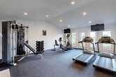 Located at 6075 Grelot Rd in Mobile, Alabama, this single-story fitness center is thoughtfully divided into two distinct workout zones, each designed to support a different style of training. The main cardio and strength room features durable carpet flooring and is equipped with Matrix treadmills, an elliptical, a stationary bike, and a multi-station cable machine accompanied by a full rack of dumbbells. Recessed lighting and vaulted ceilings lend the space an open, airy feel, while a wall-mounted flat-screen TV and French doors with views of the outdoors enhance the workout environment. A standout architectural feature of this room is a classic white-painted brick fireplace, adding a unique and warm aesthetic contrast to the fitness setting. The adjacent functional training studio offers a smooth hardwood-style floor ideal for dynamic movement and flexibility work. This room is outfitted with a multi-tier functional training rack holding medicine balls, kettlebells, and TRX suspension equipment, along with a set of stability balls and a BOSU trainer. A large wall-mounted TV and natural light from multiple windows complete the space. A dedicated entry area with a wall-mounted water fountain and restroom access adds convenience for users throughout the facility.