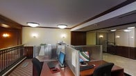 This single-floor office space combines classic architectural elegance with practical workplace functionality. The interior is defined by warm cream-colored walls accented with rich dark wood trim and crown molding, lending the environment a refined, professional character. Flush-mount ceiling light fixtures and wall-mounted sconces provide consistent, warm ambient lighting throughout the workspace.

The open-plan work area features modular workstations separated by sleek, semi-transparent cubicle partitions with horizontal slat detailing, offering employees a balance of privacy and openness. Each workstation is equipped with dual monitors and full desktop setups, reflecting a tech-forward work environment. Desks are finished in a warm wood tone, complementing the overall interior palette.

Shared office equipment, including a multifunction printer, is conveniently positioned within the open workspace area. Toward the rear of the space, glass-paneled private offices or enclosed meeting areas are visible, featuring dark wood doors and framed glass walls that allow natural light to filter through while maintaining acoustic separation. The flooring transitions between carpeted workstation areas and hard-surface flooring in adjacent zones, adding visual distinction between spaces. Overall, the office presents a well-appointed, structured environment suited for a professional services setting.