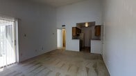 This single-floor residential space is defined by its notably high vaulted ceilings in the main living area, which lend a sense of openness and volume to the room. The living area is finished with wall-to-wall carpeting and is bathed in soft, ambient interior lighting. A wide arched passageway connects the living space to the kitchen, which features tile flooring, wood-toned upper and lower cabinetry, a refrigerator, a range, and a built-in countertop bar area — providing a practical and efficient layout for everyday use. The kitchen's design allows for a natural flow between the cooking and living spaces. A doorway visible from the main living area leads to additional rooms, including what appears to be a hallway connecting to further living quarters or a bathroom, with warm lighting visible beyond. The overall color palette is neutral throughout, with white and gray walls complementing the natural wood tones of the cabinetry. The layout is straightforward and functional, making efficient use of the available space across the single floor. The high ceilings remain a standout architectural feature that elevates the overall feel of the interior.