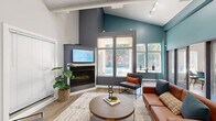 This thoughtfully designed single-floor space makes a striking first impression with its soaring vaulted ceilings and bold two-tone color palette, pairing crisp white upper walls with deep teal accents that draw the eye upward. The main living area is anchored by a sleek corner fireplace topped with a mounted flat-screen display, creating a functional and visually compelling focal point. Large multi-pane windows flood the space with natural light and offer views of the surrounding greenery, enhancing the open, airy atmosphere. Track lighting along the ceiling provides additional illumination throughout.

The living area is furnished with a warm cognac leather sofa, modern accent chairs with orange cushions, a round wood-and-metal coffee table, and a patterned area rug, all arranged to encourage comfortable gathering. Light wood-tone flooring runs throughout, complementing the contemporary aesthetic. A small service counter with dark cabinetry is visible near the entrance, adding functional utility.

Along one wall, a row of glass-paneled French doors opens into a series of private enclosed rooms, each outfitted with seating and work surfaces — ideal for focused work or meetings. The entrance area features high ceilings with framed artwork and a welcoming layout. Overall, the space balances modern design with practical versatility.