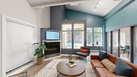 This thoughtfully designed single-floor space makes a striking first impression with its soaring vaulted ceilings and bold two-tone color palette, pairing crisp white upper walls with deep teal accents that draw the eye upward. The main living area is anchored by a sleek corner fireplace topped with a mounted flat-screen display, creating a functional and visually compelling focal point. Large multi-pane windows flood the space with natural light and offer views of the surrounding greenery, enhancing the open, airy atmosphere. Track lighting along the ceiling provides additional illumination throughout.

The living area is furnished with a warm cognac leather sofa, modern accent chairs with orange cushions, a round wood-and-metal coffee table, and a patterned area rug, all arranged to encourage comfortable gathering. Light wood-tone flooring runs throughout, complementing the contemporary aesthetic. A small service counter with dark cabinetry is visible near the entrance, adding functional utility.

Along one wall, a row of glass-paneled French doors opens into a series of private enclosed rooms, each outfitted with seating and work surfaces — ideal for focused work or meetings. The entrance area features high ceilings with framed artwork and a welcoming layout. Overall, the space balances modern design with practical versatility.