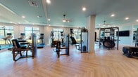 This expansive fitness center is designed to accommodate a comprehensive workout experience within a single, open-plan floor. The space is immediately distinguished by its elegant herringbone-patterned hardwood flooring, which runs throughout and lends a polished, upscale aesthetic uncommon in traditional gym settings. Tray ceilings painted in a soft teal hue add architectural interest overhead, complemented by recessed lighting and ceiling fans that maintain a bright, comfortable environment.

Large windows line the perimeter walls, flooding the interior with natural light and offering views of the surrounding greenery outside. Full-length mirrors are strategically positioned to enhance the sense of space and support proper form during training.

The equipment selection is extensive and thoughtfully arranged. A long row of treadmills runs along the window-facing wall, each unit equipped with individual screens. The cardio zone also includes ellipticals, stair climbers, and stationary bikes. The strength training area features TKO-branded cable machines, a multi-functional power rack, punching bags, kettlebells, dumbbells, and adjustable benches. A functional training zone includes a tire for conditioning drills. Glass-paneled doors lead to an adjacent room, suggesting additional amenity space nearby. Overall, the fitness center offers a clean, modern, and motivating environment for all fitness levels.
