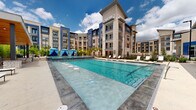 This impressive outdoor amenity space is designed to deliver a resort-style experience within a contemporary multi-story residential community. The centerpiece is a generously sized swimming pool with crystal-clear blue water, flanked by a wide pool deck outfitted with rows of sleek lounge chairs ideal for sunbathing. Submerged stepping stones add a playful design element to the pool's interior. A covered pavilion with a wood-paneled ceiling and an outdoor fireplace serves as an elegant gathering space, featuring comfortable seating arranged for relaxation and entertaining. Adjacent to the pool, a dedicated cabana zone offers vibrant blue-draped canopies for shade and privacy. A stone-accented fire pit surrounded by Adirondack-style chairs creates an inviting social atmosphere, framed by lush bamboo and tropical plantings that add greenery and natural screening. A glass-railed terrace provides additional seating with open views across the pool courtyard. The landscaping throughout incorporates ornamental grasses, palms, and native plantings, enhancing the outdoor ambiance. The surrounding building exterior blends neutral stucco, stone tower accents, and bold navy blue architectural panels, contributing a modern and polished aesthetic to the entire outdoor living environment.