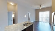 Situated at 131 E Davie Street in the heart of Raleigh, North Carolina, this single-floor residence offers a thoughtfully designed open-concept living space that combines modern aesthetics with functional comfort. The kitchen serves as a central focal point, showcasing a granite-topped island with a double stainless steel sink, dark espresso cabinetry, and elegant pendant lighting that hangs from impressively high ceilings. Rich, dark hardwood flooring flows seamlessly throughout the main living area, adding warmth and sophistication to the space. The living area is bright and airy, benefiting from natural light that filters in through a large sliding glass door dressed with vertical blinds, hinting at outdoor access. A ceiling fan overhead enhances the comfort of the living space. Beyond the kitchen, a hallway leads to a bedroom featuring carpeted flooring and a window that allows for additional natural light. The adjacent bathroom, visible from the kitchen area, continues the refined design language with matching cabinetry. The overall ambiance is clean, contemporary, and welcoming, with neutral tones and high-end finishes that create an inviting and polished living environment in one of Raleigh's most vibrant urban neighborhoods.