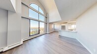 This two-storey, 2-bedroom, 3-bathroom residence in Calgary, AB offers a thoughtfully designed layout spread across two generous floors. The main level features an open-concept living, dining, and kitchen area anchored by a striking floor-to-ceiling arched window that floods the space with natural light and frames sweeping views of the surrounding cityscape and nearby water. Soaring vaulted ceilings amplify the sense of openness, while light hardwood-style flooring flows throughout. The modern kitchen is outfitted with high-gloss white cabinetry, marble-style quartz countertops, stainless steel appliances, and a peninsula island. A balcony off the main living area extends the living space outdoors with panoramic views of Calgary's skyline and natural landscape. The upper level, accessed via a bright open staircase, includes a bonus room bathed in light from the arched window below, along with both bedrooms. The primary bedroom offers generous proportions and large windows with treetop and city views. The three bathrooms are finished with clean, modern fixtures including white vanities and quartz countertops. Additional conveniences include in-unit laundry, multiple hallways for easy circulation, and an entrance foyer.