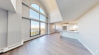 This two-storey, 2-bedroom, 3-bathroom residence in Calgary, AB offers a thoughtfully designed layout spread across two generous floors. The main level features an open-concept living, dining, and kitchen area anchored by a striking floor-to-ceiling arched window that floods the space with natural light and frames sweeping views of the surrounding cityscape and nearby water. Soaring vaulted ceilings amplify the sense of openness, while light hardwood-style flooring flows throughout. The modern kitchen is outfitted with high-gloss white cabinetry, marble-style quartz countertops, stainless steel appliances, and a peninsula island. A balcony off the main living area extends the living space outdoors with panoramic views of Calgary's skyline and natural landscape. The upper level, accessed via a bright open staircase, includes a bonus room bathed in light from the arched window below, along with both bedrooms. The primary bedroom offers generous proportions and large windows with treetop and city views. The three bathrooms are finished with clean, modern fixtures including white vanities and quartz countertops. Additional conveniences include in-unit laundry, multiple hallways for easy circulation, and an entrance foyer.
