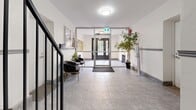 This building lobby presents a clean and functional common area designed to serve as both an entrance and transitional space for occupants. The layout encompasses a welcoming front entry vestibule with large glass-paneled doors that invite natural light into the space, complemented by flush ceiling light fixtures that ensure consistent illumination throughout.

The main lobby area features grey marble-effect tile flooring that extends seamlessly through the entry, seating area, and connecting corridor. A prominent dark metal staircase serves as a structural focal point, its bold ironwork railing contrasting elegantly against the crisp white walls. A two-toned wall treatment, divided by a horizontal grey rail, adds architectural character to the interior.

A small seating arrangement — comprising two plush dark velvet accent chairs and a round side table adorned with fresh flowers — provides a comfortable waiting area for residents and visitors. An information board mounted on the wall offers a practical community notice display. The space connects to a well-lit interior corridor through a grey-framed glass door, ensuring smooth circulation throughout the building. Overall, the lobby combines practicality with a tidy, welcoming aesthetic suited to a well-maintained residential building.