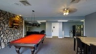 This single-floor recreational space is thoughtfully designed for leisure and entertainment. The room is anchored by a full-size pool table with a vibrant red felt surface, positioned beneath a dedicated overhead pendant light that ensures optimal visibility during play. The carpeted flooring adds warmth and comfort throughout the space.

A striking stone accent wall serves as a defining architectural feature, lending a rustic, textured contrast to the otherwise clean, blue-gray painted walls. A lounge area is situated toward the back of the room, furnished with seating ideal for relaxing between games or socializing.

Lighting is a notable element of the space, combining a decorative ceiling fan fixture with flush-mounted fluorescent panels that provide consistent, even illumination across the room. The ceiling is standard height, contributing to a comfortable, enclosed atmosphere suited for indoor recreation.

A private restroom is accessible directly from the main room, adding convenience for guests. Additional amenities such as a small refrigerator are also present, supporting extended use of the space for gatherings and events. Overall, this recreational room offers a well-equipped, inviting environment designed for social interaction and casual entertainment.