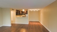 This single-floor space offers a thoughtfully designed open-concept layout that seamlessly connects the kitchen and living areas, creating a bright and functional environment. The kitchen is defined by rich, dark wood cabinetry that provides ample storage, complemented by stainless steel appliances including a microwave and range. A half-wall pass-through counter bridges the kitchen and the main living area, enhancing the sense of openness while maintaining a subtle division between the two spaces. The living area is expansive and airy, bathed in warm, ambient light from a set of track lights mounted on the ceiling, which can be directed to highlight different areas of the room. Warm-toned hardwood-style flooring runs throughout the space, adding a sense of continuity and elegance underfoot. The neutral, light-colored walls provide a versatile backdrop, making the space easy to personalize. Clean white baseboards frame the floors with a crisp finish. The overall ambiance is modern yet inviting, with a cohesive design that balances practicality and style, making it well-suited for both everyday living and entertaining.