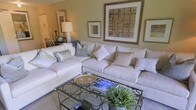 This single-floor apartment at 7915 Chrisford Place, Unit G, offers a well-appointed living space designed with comfort and style in mind. The living room serves as the focal point of the home, showcasing a large, plush sectional sofa in a soft cream tone, adorned with an assortment of decorative pillows in varying textures and subtle patterns. The neutral color palette of warm beiges and soft whites creates a calm, inviting atmosphere throughout.

A glass-top coffee table with an ornate metal base anchors the seating area, adding a refined decorative touch while maintaining visual openness. The walls are tastefully decorated with a curated selection of framed artwork, including a large statement piece with earthy, abstract tones that complements the overall color scheme. A classic table lamp provides warm ambient lighting, enhancing the cozy feel of the room.

Lush greenery in the form of a potted plant introduces a natural element, adding vibrancy to the neutral surroundings. A glimpse into the adjoining space reveals additional furnishings, suggesting a thoughtfully laid-out floor plan. Overall, the space conveys a warm, sophisticated ambiance suited for comfortable everyday living.