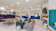 Located at 2301 E Tenth St in Greenville, North Carolina, this generously sized single-floor amenity space is designed to foster community and recreation. The layout flows seamlessly from one zone to the next, all unified by polished terrazzo flooring that runs throughout. Bold cobalt blue accent walls paired with vibrant artwork and colorful furnishings give the space an energetic, modern character.

The game and lounge area anchors one end of the space, featuring ping-pong tables, plush sectional sofas with accent pillows, and casual seating arrangements ideal for social gatherings. A decorative fireplace with a tiled surround and a wall-mounted TV creates a cozy focal point nearby. Elegant chandelier lighting adds warmth and sophistication above a long communal dining table surrounded by bright teal bar stools.

A kitchenette offers granite countertops, white cabinetry, a stainless steel sink, and a classic subway tile backsplash, providing functional support for events and everyday use. Transitional hallways connect quieter collaborative nooks furnished with built-in banquette seating, high-top tables, and distinctive sculptural pendant lights. Large windows and glass doors flood the space with natural light while offering views of the surrounding outdoor environment, enhancing the open and inviting ambiance throughout.