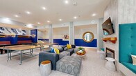 Located at 2301 E Tenth St in Greenville, North Carolina, this generously sized single-floor amenity space is designed to foster community and recreation. The layout flows seamlessly from one zone to the next, all unified by polished terrazzo flooring that runs throughout. Bold cobalt blue accent walls paired with vibrant artwork and colorful furnishings give the space an energetic, modern character.

The game and lounge area anchors one end of the space, featuring ping-pong tables, plush sectional sofas with accent pillows, and casual seating arrangements ideal for social gatherings. A decorative fireplace with a tiled surround and a wall-mounted TV creates a cozy focal point nearby. Elegant chandelier lighting adds warmth and sophistication above a long communal dining table surrounded by bright teal bar stools.

A kitchenette offers granite countertops, white cabinetry, a stainless steel sink, and a classic subway tile backsplash, providing functional support for events and everyday use. Transitional hallways connect quieter collaborative nooks furnished with built-in banquette seating, high-top tables, and distinctive sculptural pendant lights. Large windows and glass doors flood the space with natural light while offering views of the surrounding outdoor environment, enhancing the open and inviting ambiance throughout.