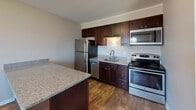 Located at 5 Cinema Dr in Lansing, NY, this single-floor unit offers a thoughtfully designed layout with a combination of functional and comfortable living spaces. The open-concept kitchen is a standout feature, equipped with stainless steel appliances including a refrigerator, dishwasher, electric range, and microwave. Rich dark-wood cabinetry provides ample storage, while granite-style countertops and a stainless steel sink add a polished finish. Wood-style flooring grounds the kitchen area and flows into the adjoining dining space.

The living area is generously sized and finished with plush carpeting in a neutral tone. A sliding glass door opens to a private balcony, allowing natural light to fill the space and providing outdoor views of the surrounding area.

The bedrooms are carpeted and feature neutral walls that lend a calm, versatile atmosphere. One bedroom includes multiple windows that invite natural light, along with a built-in shelving unit and closet. Another bedroom offers a double-door closet for convenient storage.

The bathroom is finished with tile flooring, a warm wood-toned vanity with a white countertop, an illuminated mirror, and a recessed medicine cabinet, completing this well-maintained and move-in ready space.