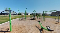 This outdoor community space centers around a well-equipped playground designed to encourage active play and physical development. The play area is surfaced with a generous layer of wood chip mulch, providing a safe and impact-absorbing ground cover beneath and around all equipment. The playground features a collection of green metal climbing structures, including rope-and-frame climbing towers that rise to varying heights, offering children multiple challenges and vantage points. A seesaw and additional activity stations round out the equipment selection, ensuring a variety of play experiences. A curved archway structure is visible at the perimeter, adding a distinctive architectural element to the space. The playground is bordered by a curbed pathway that defines the boundary between the play surface and the surrounding landscaped lawn. The open green space adjacent to the play area provides ample room for open-ended outdoor activities. Residential homes are visible in the background, situating this playground as a neighborhood amenity. The setting benefits from abundant natural light and open sky, creating an inviting and energetic outdoor environment suited for recreational use throughout the day.