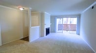 This single-floor residence offers an open and airy living area defined by thoughtful architectural details and a warm, neutral palette. The main living space is anchored by a classic fireplace with a white mantel, serving as a natural focal point. Flanking the fireplace is a decorative column with crown molding that adds architectural character and subtly defines the transition between areas. A pass-through window or built-in niche is integrated into the partial wall, enhancing the open feel while maintaining a sense of structure. Plush wall-to-wall carpeting extends throughout, contributing to a comfortable, quiet atmosphere. Crown molding trims the ceilings, lending a polished, finished look to the space. A textured ceiling adds subtle visual interest overhead. Natural light floods the interior through expansive sliding glass doors that lead directly to a private balcony with a wooden railing, offering outdoor views and easy indoor-outdoor access. The overall ambiance is bright, clean, and welcoming, with a layout that balances openness with defined living zones.
