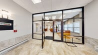 Located at 311 North Ottawa Street in Joliet, Illinois, this single-floor facility offers a generous and well-appointed layout designed for both comfort and functionality. Entry is made through sleek, dark-framed glass doors that open into a bright reception area, complete with a warm wood reception desk, comfortable leather seating, and large windows fitted with blinds that invite natural light throughout.

The open lounge area is an inviting space featuring polished stone-look tile flooring, ceiling fans, recessed skylights, and tasteful wall sconces. A sculptural white art installation serves as a striking focal point, while a bar-height communal table with wooden chairs provides a relaxed gathering spot. Framed artwork and a decorative wall clock add character to the warm, neutral-toned interior.

A café area offers round white tables and wooden chairs arranged around a stylish service counter adorned with a hexagonal tile facade and topped with a light quartz surface. Light wood-look flooring and bright panel lighting create a clean, modern atmosphere ideal for casual dining or socializing.

The dedicated exercise room is generously sized and well-equipped, featuring rows of treadmills, ellipticals, stationary bikes, and a variety of cable strength machines. Large windows line the walls, large mirrors amplify the sense of space, and ceiling fans ensure comfort during workouts. An adjacent area provides locker storage and a wall-mounted television, completing a comprehensive fitness facility.