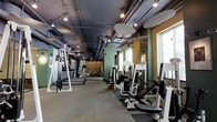 This fitness center offers a spacious, single-floor layout designed to accommodate a comprehensive workout experience. The space features an industrial aesthetic, with exposed ductwork, pipes, and ceiling infrastructure painted in a deep charcoal tone, contrasting with sage green and warm ochre accent walls that lend the environment a modern, energetic character. Track lighting and pendant fixtures illuminate the floor evenly, ensuring excellent visibility throughout the facility. The main workout area is generously proportioned, with an open floor plan that allows for comfortable spacing between equipment. A robust selection of strength training machines lines both sides of the space, including cable systems, chest press, leg press, and various resistance machines suited to a full-body routine. Cardio equipment, including treadmills, is arranged along one side of the floor. Rubberized flooring beneath the weight machines provides both safety and durability. Large windows along one wall invite natural light into the space, enhancing the overall atmosphere. Mirrored wall sections further amplify the sense of openness while supporting proper form during training sessions. The layout is thoughtfully organized to ensure smooth traffic flow between equipment zones.
