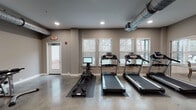 This fitness center in Lakeland, Tennessee offers a comprehensive single-floor workout facility designed to accommodate a wide range of exercise routines. The space features polished concrete flooring throughout, lending it an industrial-modern aesthetic that is both durable and visually clean. Exposed silver ductwork runs across the ceiling, complementing the neutral gray walls and recessed lighting to create a contemporary, functional atmosphere.

The cardio zone is lined along a windowed wall, where multiple treadmills and an elliptical machine are positioned to allow natural light from the slatted-blind windows to filter in, providing views of the surrounding wooded landscape. A rowing machine with a digital monitor rounds out the cardio offerings in this area.

The strength and functional training zone is well-appointed with a Schwinn air bike, adjustable weight benches, a dumbbell rack, medicine balls, resistance bands, and a TRX suspension training system. Wall-mounted flat-screen televisions are positioned throughout the space to keep users engaged during workouts.

A distinctive floor-to-ceiling glass partition wall separates the fitness center from an adjacent community lounge, maintaining visual openness while defining each space. A private door provides direct exterior access, adding convenience for users entering from outside.