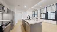 Located at 424 S Broadway in the heart of Downtown Los Angeles, Unit 404 is a thoughtfully designed studio offering an efficient and modern living layout on a single floor. The open main living area is anchored by large, black-framed multi-pane windows dressed with dual-shade roller blinds, flooding the space with natural light while offering a nod to the building's urban character. Light-toned hardwood flooring runs throughout, lending warmth and continuity across the entire unit.

The kitchen is a standout feature, designed with clean, contemporary lines. A sleek gray island with a light countertop serves as both a prep space and a casual dining surface. Behind it, light-toned cabinetry pairs with a classic white subway tile backsplash, and stainless steel appliances — including a refrigerator, microwave, and range — complete the functional setup. Recessed lighting illuminates the kitchen and living areas cleanly overhead.

The bathroom is finished in crisp white, featuring a combination tub and shower with white tile surround, a wall-mounted sink, and an accent of mosaic tile detailing that adds a subtle decorative touch. A sliding glass door provides a sense of openness to the adjacent space. The overall design of the unit is clean, minimal, and well-suited to urban living in one of Los Angeles' most vibrant neighborhoods.
