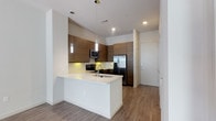 This single-floor residence offers a thoughtfully designed 1-bedroom, 1-bathroom layout that maximizes functionality and style. The open-concept kitchen serves as a central highlight, featuring flat-panel cabinetry in a warm wood-grain finish that contrasts elegantly against crisp white quartz countertops. A peninsula with an integrated sink provides additional prep space and casual dining potential. Stainless steel appliances, including a French door refrigerator, microwave, and range, lend a polished, modern aesthetic. Pendant lighting suspended above the peninsula adds a decorative touch while providing task illumination, complemented by recessed ceiling lights throughout. The kitchen flows seamlessly into the adjacent living areas, creating an open and connected feel. Wood-look plank flooring extends throughout the main living space, offering both durability and a warm, contemporary look. White walls and clean-lined finishes contribute to a bright, neutral palette that is easy to personalize. A private entry door is visible from the kitchen, providing a clear and practical layout. Overall, the space presents a clean, modern aesthetic well-suited for comfortable everyday living.
