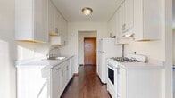 This single-floor residential unit offers a well-organized layout with distinct living spaces, each finished with practical and clean design elements. The galley-style kitchen is a standout feature, lined on both sides with white shaker cabinetry equipped with sleek bar-pull hardware, offering generous storage from floor to ceiling. A stainless steel sink sits along one wall, while a white gas range and refrigerator occupy the opposite side, all set above rich, dark hardwood flooring. The kitchen opens toward a bright dining area, illuminated by a pendant light fixture and a wood-framed window that draws in natural light. The living area is spacious and airy, featuring the same hardwood flooring, large wood-trimmed windows that allow abundant natural light to fill the room, baseboard heating, and a wall-mounted air conditioning unit. The bedroom is carpeted for comfort, with natural light filtering through a wood-framed window and baseboard heating along the wall. The full bathroom features classic pink mosaic tile flooring, white subway tile walls, a white vanity with mirror, and a combined tub and shower, offering a retro yet functional aesthetic throughout.