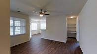 Located at 4625 Millbrook Green Dr in Raleigh, NC, this single-floor residence offers a welcoming and functional living environment defined by clean lines and thoughtful design. The main living area showcases rich, dark hardwood flooring that runs throughout, creating a warm and cohesive aesthetic. A ceiling fan with a light fixture anchors the living space, adding both comfort and charm. Natural light flows generously into the space through multiple windows dressed with horizontal blinds, including a distinctive bay window area that adds architectural interest and serves as an inviting dining nook, accented by a pendant light overhead. The open layout connects the living and dining areas seamlessly, lending a spacious and unobstructed feel to the interior. Adjacent to the main living space, a built-in shelving unit provides practical storage with a refined, finished look, while nearby countertop surfaces hint at a well-appointed kitchen or utility area with granite-style detailing. The neutral wall tones throughout create a versatile backdrop, making the space adaptable to a variety of interior styles. Overall, this residence balances everyday practicality with tasteful design elements, making it a comfortable and attractive home in the Raleigh area.