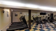 This single-floor fitness center offers a practical and functional workout environment designed to accommodate a variety of exercise needs. The space features a distinctive blue and yellow checkered floor that adds a lively visual character to the room. Overhead, fluorescent strip lighting runs along the ceiling perimeter, providing bright, even illumination throughout the area. A textured ceiling and structural support columns are characteristic architectural elements of the space.

The fitness center is equipped with multiple cardio machines, including treadmills and stationary bikes, positioned to allow comfortable use and spacing between equipment. A dedicated strength training area includes a dumbbell rack with an organized selection of free weights, complemented by additional resistance and weight machines. A wall-mounted flat-screen television is positioned for visibility during workouts, adding an entertainment element to the exercise experience.

Motivational fitness posters and workout illustration charts are displayed along the walls, contributing to an encouraging atmosphere. The overall layout is efficient and thoughtfully arranged, making the most of the available floor space while ensuring a clear and navigable workout area. This fitness center provides residents or guests with a convenient, all-in-one facility for maintaining an active lifestyle.