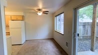 This single-floor space presents a straightforward and functional layout designed for comfortable everyday living. The main living area is finished with wall-to-wall carpeting that adds warmth underfoot, while neutral-toned walls provide a clean and versatile backdrop. A ceiling fan with an integrated light fixture is centrally positioned overhead, offering both air circulation and ambient lighting throughout the room. Natural light enters through a standard window, providing views to an outdoor fenced area and keeping the space bright. A sliding glass door offers convenient access to the exterior, enhancing the connection between indoor and outdoor areas. A floor-level vent indicates a built-in climate control system for year-round comfort. The space also features a partial view of an adjacent area, suggesting additional functional zones nearby. Overall, the space is modest in scale yet efficiently arranged, with practical finishes and essential features that make it a well-suited environment for a variety of uses.