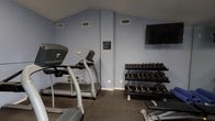 Located at 2945 Layfair Dr in Flowood, Mississippi, this dedicated fitness center offers a well-equipped, single-floor workout facility designed to support a full range of exercise routines. The space features vaulted ceilings that lend an open, airy feel, and is finished with durable rubber flooring suitable for high-traffic athletic use throughout.

The cardio area is equipped with multiple treadmills and an elliptical machine, providing ample options for cardiovascular training. A stationary upright bike occupies its own section of the room, positioned near natural light from windows fitted with horizontal blinds. A wall-mounted flat-screen television is positioned for easy viewing during workouts.

The strength training area includes a multi-tiered dumbbell rack with a comprehensive range of free weights, a kettlebell rack stocked with various sizes and weights, a weight bench, and yoga/exercise mats for floor-based training. Full-length glass mirrors line one wall, expanding the sense of space and allowing users to monitor their form.

Practical amenities include a water cooler, waste receptacle, a posted heart rate target chart, and clearly displayed fitness rules. The facility is also equipped with a fire extinguisher for safety. Natural light enters through multiple windows, complementing the perimeter lighting and contributing to an inviting, functional atmosphere well-suited for consistent, effective workouts.