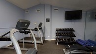 Located at 2945 Layfair Dr in Flowood, Mississippi, this dedicated fitness center offers a well-equipped, single-floor workout facility designed to support a full range of exercise routines. The space features vaulted ceilings that lend an open, airy feel, and is finished with durable rubber flooring suitable for high-traffic athletic use throughout.

The cardio area is equipped with multiple treadmills and an elliptical machine, providing ample options for cardiovascular training. A stationary upright bike occupies its own section of the room, positioned near natural light from windows fitted with horizontal blinds. A wall-mounted flat-screen television is positioned for easy viewing during workouts.

The strength training area includes a multi-tiered dumbbell rack with a comprehensive range of free weights, a kettlebell rack stocked with various sizes and weights, a weight bench, and yoga/exercise mats for floor-based training. Full-length glass mirrors line one wall, expanding the sense of space and allowing users to monitor their form.

Practical amenities include a water cooler, waste receptacle, a posted heart rate target chart, and clearly displayed fitness rules. The facility is also equipped with a fire extinguisher for safety. Natural light enters through multiple windows, complementing the perimeter lighting and contributing to an inviting, functional atmosphere well-suited for consistent, effective workouts.
