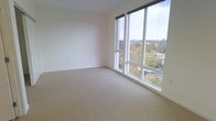 This single-floor space is defined by its clean, modern aesthetic and an abundance of natural light. The standout feature is a dramatic set of floor-to-ceiling windows that span an entire corner of the room, framing expansive views of the surrounding neighborhood and its lush, mature tree canopy. The elevated vantage point provides a sense of openness and connection to the outdoors, with glimpses of street-level activity visible below.

The interior is finished with neutral, wall-to-wall carpeting in a warm beige tone, contributing to a calm and understated atmosphere. The smooth, white walls are clean and minimal, offering a versatile canvas that suits a variety of uses and furnishing styles. The overall layout feels open and generously proportioned, with the corner window configuration enhancing the sense of spaciousness.

The design prioritizes light and views, making this a space that feels airy throughout the day. The modern window framing and simple, uncluttered finishes lend the space a contemporary character. Whether used as a residential living area, a home office, or a flexible multi-purpose room, the combination of natural light, elevated views, and neutral finishes makes this a highly functional and visually appealing environment.