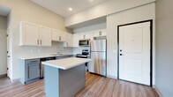This single-floor unit showcases a contemporary design with a neutral color palette and durable luxury vinyl plank flooring throughout the main living areas. The kitchen serves as a central highlight, featuring two-tone cabinetry with white upper cabinets and slate-gray lower cabinets, all accented by sleek black hardware. A freestanding center island with a crisp white countertop provides additional workspace and casual seating. Stainless steel appliances — including a refrigerator, electric range, microwave, and dishwasher — complete the functional and modern kitchen setup.

The bedrooms are carpeted for comfort, finished with neutral gray walls and white two-panel doors with black hardware. One bedroom benefits from a large window offering views of mature trees and greenery outdoors, bringing in natural light. A dedicated laundry room houses a full-size top-loading washer and dryer with overhead wire shelving for storage convenience.

The bathroom features a gray vanity with a white countertop, a large mirror, and luxury vinyl plank flooring, maintaining the consistent modern aesthetic found throughout the unit. Overall, the space presents a clean, move-in ready environment with cohesive design elements.