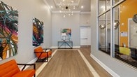 This stylish lobby space makes a strong first impression with its soaring ceilings and open, airy layout. The flooring throughout is a warm wood-look plank tile accented by a contrasting inlay border, adding architectural interest underfoot. A striking sputnik-style chandelier anchors the main reception area, complemented by recessed lighting and a coffered ceiling detail that adds depth and sophistication.

Large floor-to-ceiling windows with black metal framing line one side of the lobby, allowing generous amounts of natural light to pour in while offering views to the exterior. Double glass entry doors with sheer roller shades provide a bright, welcoming point of entry. Vibrant, large-format abstract artwork adorns the walls, adding bold pops of color against the neutral gray palette. Modern orange accent chairs with sleek black metal frames provide seating and echo the energy of the artwork.

A passenger elevator with brushed metal doors is conveniently accessible from the lobby corridor, alongside additional doorways leading to connected areas of the building. The hallways maintain the same cohesive aesthetic with consistent flooring and high ceilings. Overall, the space balances contemporary design with functional flow, creating a polished and inviting environment suited for a professional or multi-use commercial setting.
