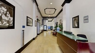 Located at 501-517 S Spring St in the heart of downtown Los Angeles, this lobby hallway exudes classic architectural character with a refined, historic ambiance. The space stretches along a generous corridor layout, featuring light-toned marble-style tile flooring that reflects the well-lit interior and contributes to an open, airy feel.

Architectural detailing is a defining feature throughout, with bold dark crown molding, coffered ceiling accents, and decorative pendant lighting that draw the eye upward and add a sense of grandeur. High ceilings amplify the spaciousness of the corridor, while white walls trimmed in deep espresso tones create a sharp, elegant contrast.

A wood-paneled reception desk with a green glass countertop serves as a central focal point, providing a welcoming point of contact for visitors. Vintage-style seating areas—including a richly upholstered red sofa, ornate wooden chairs, and a marble-top coffee table atop a zebra-print rug—offer comfortable gathering spots with old-world charm. Lush potted palms add organic warmth to the setting.

Historic framed photographs and artwork line the walls, reinforcing the building's storied character. Practical amenities, including vending machines and elevator access, are seamlessly integrated at the rear of the hallway, balancing classic aesthetics with modern convenience.