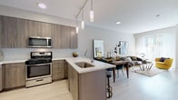 This single-floor residence at 300 Parkway Dr in Lincolnshire, Illinois, presents a thoughtfully designed open-concept layout with contemporary finishes throughout. The kitchen serves as a central feature, equipped with stainless steel appliances including a gas range, over-the-range microwave, French door refrigerator with a built-in wine rack, and dishwasher. Warm-toned wood-grain cabinetry with sleek hardware pairs with white quartz countertops and a subtle tile backsplash. A peninsula with a single-basin undermount sink and pendant lighting above provides additional prep space and casual seating, flowing naturally into the dining and living areas.

The living room is bright and welcoming, benefiting from large windows that allow ample natural light and connect to the open floor plan. Light wood-style flooring runs throughout the main living areas, contributing to a clean, cohesive aesthetic.

The bedroom features carpeted flooring, generous closet space, large windows with sheer curtains, and a bold accent wall that adds visual interest. The bathroom is finished with large-format tile flooring, a wood-grain vanity with a white countertop, a wide mirror, and a glass-enclosed walk-in shower, completing this well-appointed residence.